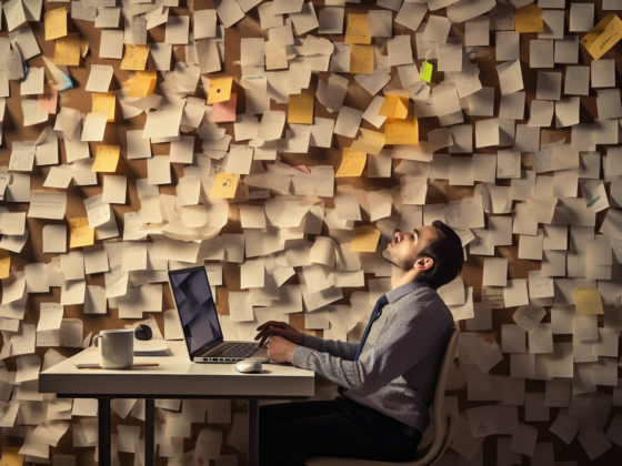 un homme est assis devant son ordi et a l'aise épuisé. En fond un énorme mur avec plein de post-its dessus.