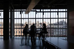 Trois personnes sont assises autour d'une table, image en contre-jour donc très sombre. Le fond est une grande vitre donnant sur le haut d'un immeuble en brique rouge