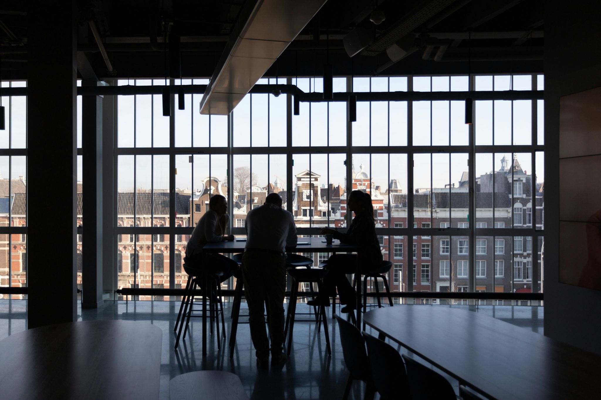 Trois personnes sont assises autour d'une table, image en contre-jour donc très sombre. Le fond est une grande vitre donnant sur le haut d'un immeuble en brique rouge